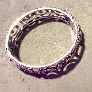 May include: A silver bangle bracelet with an intricate, swirling design. The bracelet is open and has a smooth, polished finish.