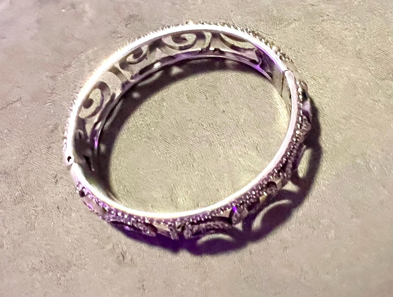 May include: A silver bangle bracelet with an intricate, swirling design. The bracelet is open and has a smooth, polished finish.
