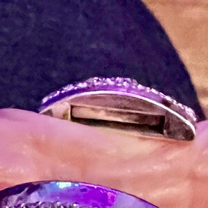 May include: A silver ring with a row of small, clear stones around the band. The ring is open on one side and has a small gap in the band.