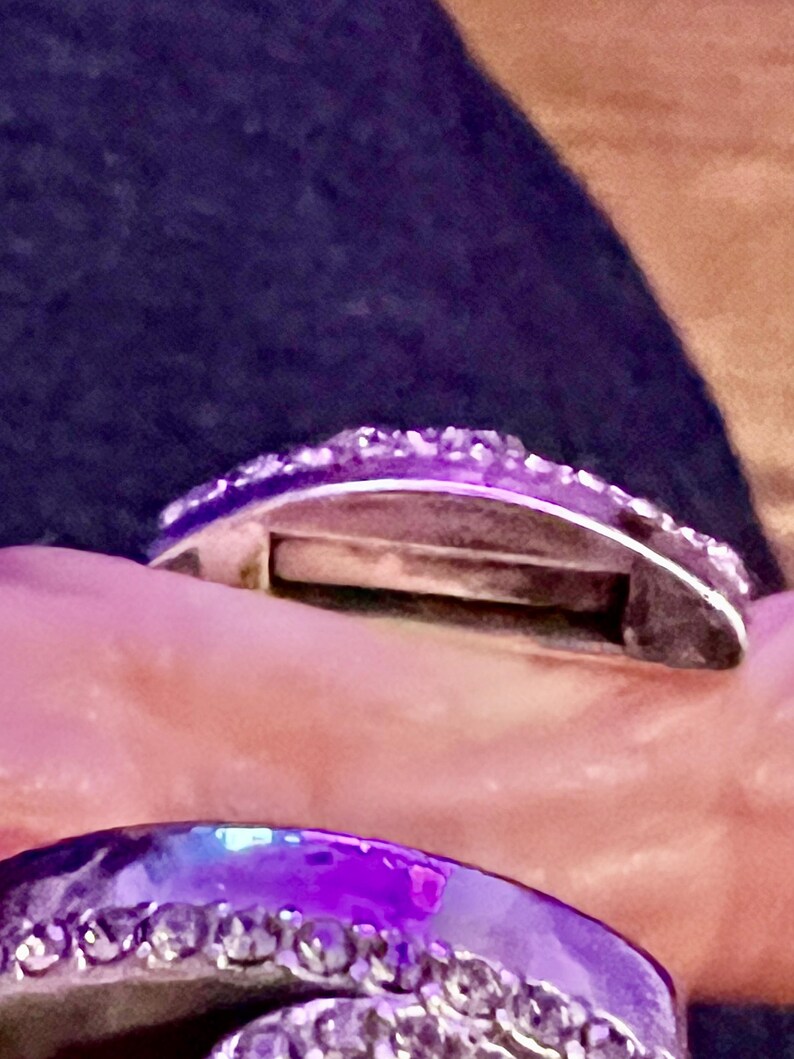 May include: A silver ring with a row of small, clear stones around the band. The ring is open on one side and has a small gap in the band.