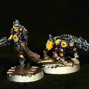 Puede incluir: Una figura en miniatura de un soldado futurista con un abrigo largo marrón con detalles amarillos que sostiene una pistola, junto a una figura de perro robótico con armadura negra y amarilla, ambas sobre bases circulares metálicas texturizadas.