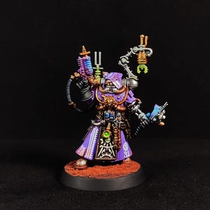 Op de afbeelding: Een geschilderd miniatuurfiguur in een paarse gewaad met capuchon en metalen gouden en zilveren mechanische details. Het figuur houdt een paars plasmapistool vast en heeft een mechanische arm met een groene klauw, een schedel-embleem en een gloeiende groene bol.