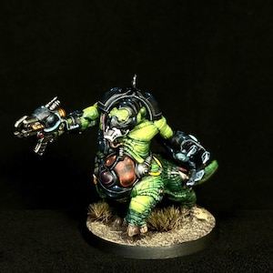Könnte beinhalten: Eine bemalte Miniaturfigur eines grünen, reptilienartigen Wesens mit muskulösem Körperbau, das eine dunkle Metallrüstung und eine weiße Gesichtsmaske trägt. Die Figur hält eine mechanische Waffe und hat ein großes, dunkles, kreisförmiges Brustteil.
