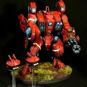 May include: A red robotic mech figure with white panel markings and a large multi-barreled cannon on its left arm. The mech features a central circular emblem, shoulder-mounted sensors, and stands on a round base with two smaller red drones.