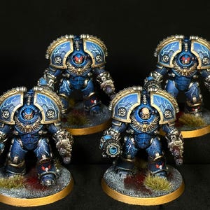 May include: Four miniature armored figures in blue power armor with gold trim and white lightning bolt patterns. Each figure features a helmeted or bare head, a chest eagle emblem, and heavy weaponry mounted on the arms, standing on circular bases.