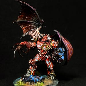 Könnte beinhalten: Eine bemalte Miniaturfigur eines geflügelten Dämonenwesens mit roter Rüstung, blauen Energieadern und einem goldenen Sonnenemblem auf der Brust. Die Figur hat große dunkle Flügel, scharfe Krallen und steht auf einem runden Sockel mit Grasbüscheln.