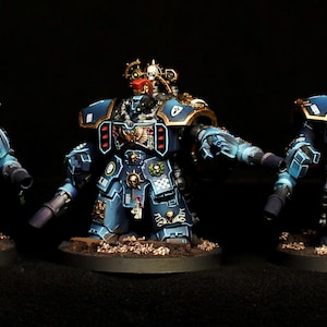 May include: Three miniature armored figures painted in shades of blue with gold trim and red accents. Each figure features a bulky, mechanical design with shoulder plates, chest emblems, and integrated weaponry on their arms, standing on circular bases.