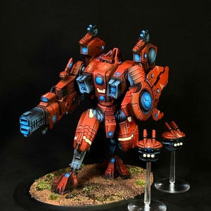 May include: A red robotic figurine with blue glowing accents and a large cannon on its right arm. The figure stands on a textured base with two smaller floating drone units on clear stands, featuring mechanical armor plating and intricate panel lines.
