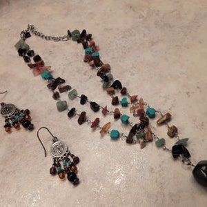 K&ouml;nnte beinhalten: Ein Halsketten- und Ohrring-Set. Die Halskette besteht aus einer silbernen Kette mit mehreren Str&auml;ngen aus farbigen, unregelm&auml;&szlig;ig geformten Steinen in T&uuml;rkis, Braun, Schwarz und Gr&uuml;n. Die Ohrringe haben ein &auml;hnliches Design mit einer silbernen Scheibe und h&auml;ngenden Steinen.