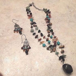 K&ouml;nnte beinhalten: Ein Halsketten- und Ohrring-Set mit einer Vielzahl farbiger Steinperlen. Die Halskette hat eine silberne Kette und einen gro&szlig;en, dunklen Steinanh&auml;nger. Die Ohrringe haben ein silbernes Design mit kleinen Perlen.