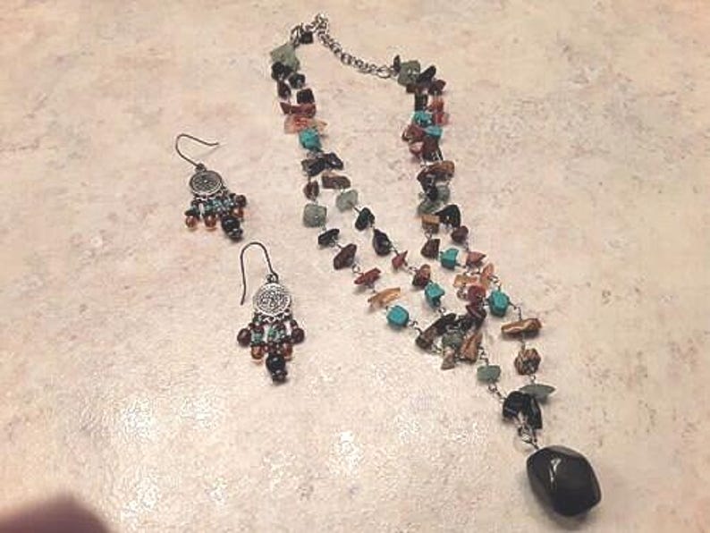 K&ouml;nnte beinhalten: Ein Halsketten- und Ohrring-Set mit einer Vielzahl farbiger Steinperlen. Die Halskette hat eine silberne Kette und einen gro&szlig;en, dunklen Steinanh&auml;nger. Die Ohrringe haben ein silbernes Design mit kleinen Perlen.