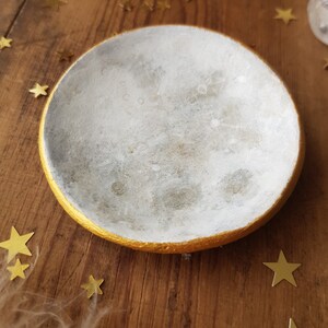 Peut inclure: Un plat en céramique blanche avec un bord doré, en forme de croissant de lune. Le plat est posé sur une surface en bois avec des étoiles dorées éparpillées autour.