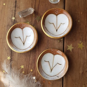 May include: Three small ceramic dishes with a brown and gold rim. Each dish features a hand-painted white owl with closed eyes. The owls are painted in a whimsical style with brown and gold accents.