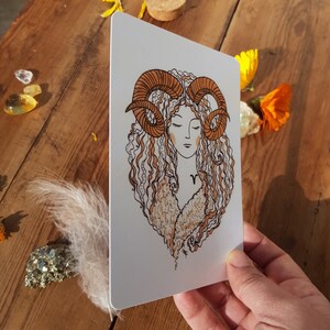 Puede incluir: Una tarjeta blanca con una ilustraci&oacute;n dibujada a mano de una mujer con cabello casta&ntilde;o rizado y cuernos de carnero. La mujer lleva una camisa blanca con un cuello de piel marr&oacute;n y un peque&ntilde;o s&iacute;mbolo 'V' en su pecho.