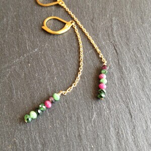 Könnte beinhalten: Goldfarbene Ohrhänger mit Klappbügelverschluss. Jeder Ohrring hat eine feine Kette, an der eine Ansammlung kleiner, runder Perlen in Grün, Rosa und Dunkelrot hängt. Die Ohrringe werden auf einer dunkelgrauen Oberfläche gezeigt.