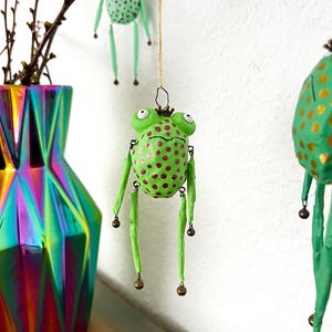 May include: Handmade green frog ornament with red polka dots and dangling legs. The frog has a small crown and is suspended by a gold string. A colorful geometric vase and branches are in the background. The frog is a whimsical decoration.