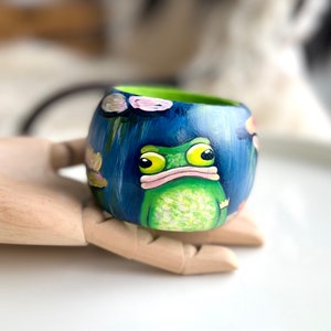 Könnte beinhalten: Ein handbemaltes Holz-Armband mit einem Design aus grünem Frosch und blauen Seerosen. Der Frosch hat große gelbe Augen und einen rosa Mund.