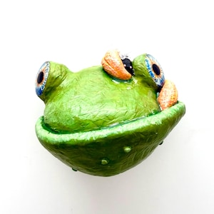 Könnte beinhalten: Handgefertigte Froschskulptur mit einem leuchtend grünen Kopf und strukturierten Details. Der Frosch hat große Augen mit blauen Iris und orangefarbenen Akzenten. Die Skulptur steht vor einem weißen Hintergrund.