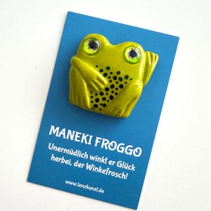Könnte beinhalten: Eine lindgrüne Froschfigur mit dunkelgrünen Punkten und großen, detaillierten Augen, befestigt auf einer blauen Präsentationskarte. Die Karte zeigt den Text MANEKI FROGGO, Unermüdlich winkt er Glück herbei, der Winkefrosch! und www.lenzkunzt.de in Weiß.
