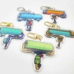 May include: Five colourful paint roller keychain charms with gold key rings. The charms are made of clear acrylic with a pixelated design. The paint rollers are blue, green, orange, and two shades of green.