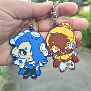 Pode incluir: Dois chaveiros coloridos de personagens de desenhos animados. Um chaveiro é azul e branco com um design amarelo e preto. O outro chaveiro é marrom e amarelo com um design branco e preto.