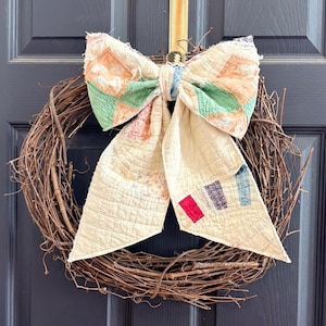 Puede incluir: Una corona rústica de vid adornada con un gran lazo hecho de una colcha vintage. El lazo presenta parches de color naranja, verde y azul, añadiendo un toque de color a la corona natural. Un encantador artículo de decoración del hogar hecho a mano.