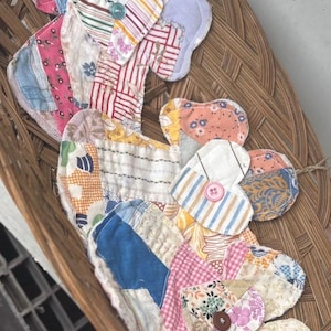 Puede incluir: Dos almohadillas de tela construidas con varios parches acolchados en colores y patrones variados, incluyendo rosa, azul, amarillo y blanco. Cada almohadilla presenta una secci&oacute;n superior en forma de coraz&oacute;n asegurada con un peque&ntilde;o bot&oacute;n de pl&aacute;stico azul, rosa o marr&oacute;n.