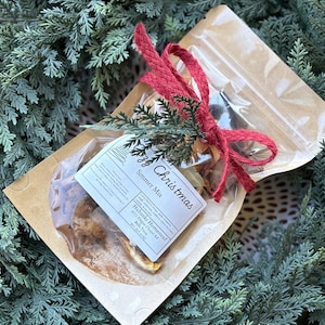 Könnte beinhalten: Ein klarer Zellophanbeutel mit einer Simmer-Mischung, gebunden mit einem roten Band und Zweigen. Der Beutel befindet sich in einem braunen Papierumschlag. Das Etikett lautet "Cozy Christmas Simmer Mix".