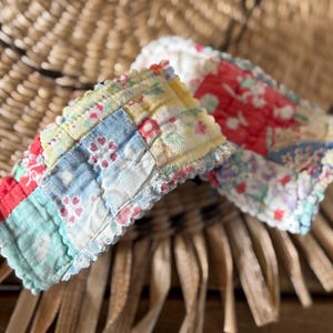 May include: Two rectangular fabric hair clips featuring a quilted patchwork design. The clips are covered in soft cotton fabric with a variety of floral and geometric patterns in shades of yellow, red, blue, green, and white, finished with scalloped edges.