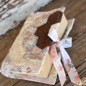 May include: A quilted book cover with a floral pattern in pastel colors. The cover features a brown applique and is tied with a ribbon printed with pink flowers and green leaves. The book is resting on a weathered wooden surface.