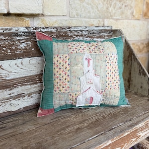 May include: A square quilted throw pillow featuring a central white fabric applique of a church building with a red cross. The pillow is constructed from patchwork fabric squares in floral patterns and solid teal green borders with visible stitching.