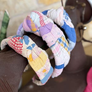 May include: Two knotted headbands crafted from multicolored patchwork fabric. The headbands feature a variety of vintage-style patterns including floral, plaid, and gingham prints in shades of pink, blue, yellow, red, and white with visible stitching.