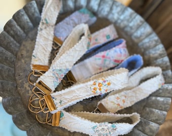 Puede incluir: Una colección de llaveros de tela para la muñeca con herrajes dorados, expuestos en una bandeja de metal galvanizado. Los llaveros presentan varios diseños de tela floral y estampada en blanco, azul y amarillo.