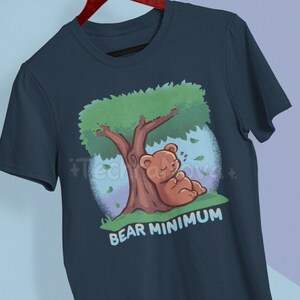 Peut inclure: Un t-shirt bleu marine avec un ours de dessin animé dormant sous un arbre vert. Le texte "BEAR MINIMUM" est imprimé sous l'ours.