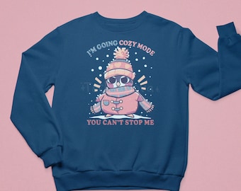 Può includere: Una felpa blu navy con un pinguino dei cartoni animati che indossa un cappello e una sciarpa rosa. Il testo "I'm going cozy mode you can't stop me" è stampato sulla felpa.