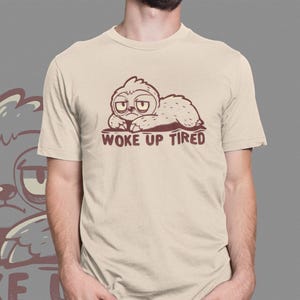 May include: Beige t-shirt featuring a brown cartoon sloth with the text "WOKE UP TIRED". The sloth is lying down with a sleepy expression. The shirt has short sleeves and a crew neck.
