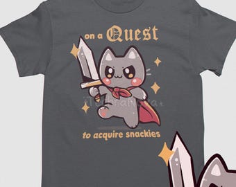 May include: A dark gray t-shirt with a cartoon gray cat holding a sword and wearing a red cape. The text on the shirt reads "on a Quest to acquire snackies".