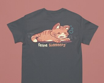 May include: A charcoal grey t-shirt with a cartoon illustration of an orange tabby cat with closed eyes and a single tear. The cat is lying on its side with its paws outstretched. The text "feline sleeeeeepy" is printed below the cat.
