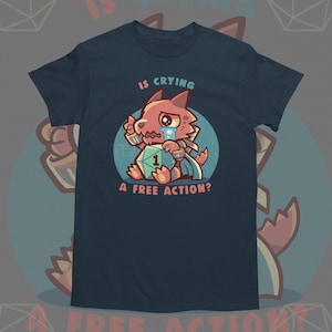 May include: Navy blue t-shirt featuring a cartoon character with a sad expression, holding a die with the number 1. The text on the shirt reads "IS CRYING A FREE ACTION?" in a playful font. The design is set in a blue circle.