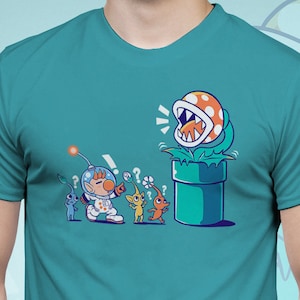 May include: Turquoise blue unisex t-shirt with a cartoon design. The illustration depicts a character in a spacesuit confronting a Piranha Plant from a green pipe, with other characters. The text "Art by TECHRANOVA" is at the bottom.