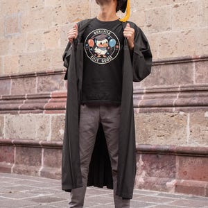 Może przedstawiać: Osoba w czarnej szacie i czapce absolwenta, trzymająca czarny t-shirt z białym kołem graficznym, na kt&oacute;rym widnieje napis "Qualified Silly Goose" z kresk&oacute;wkową gęsią w czapce absolwenta trzymającą balony.