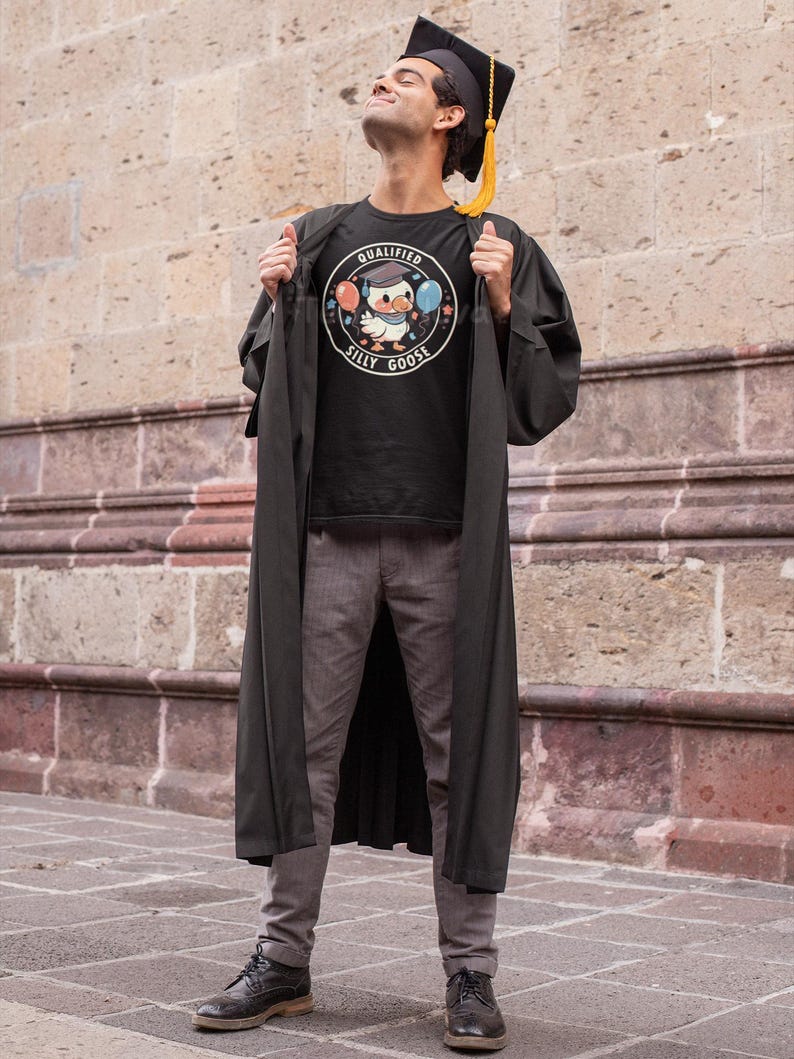 Może przedstawiać: Osoba w czarnej szacie i czapce absolwenta, trzymająca czarny t-shirt z białym kołem graficznym, na kt&oacute;rym widnieje napis "Qualified Silly Goose" z kresk&oacute;wkową gęsią w czapce absolwenta trzymającą balony.