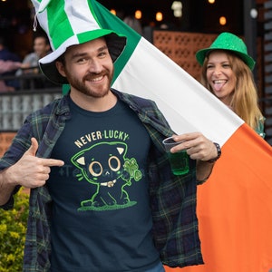 May include: A man wearing a green and white hat and a blue t-shirt with a black cat graphic and the text "Never Lucky". He is holding a green drink in a plastic cup and is standing in front of an Irish flag.