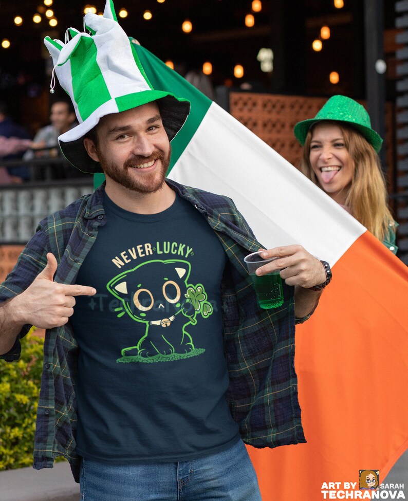May include: A man wearing a green and white hat and a blue t-shirt with a black cat graphic and the text "Never Lucky". He is holding a green drink in a plastic cup and is standing in front of an Irish flag.