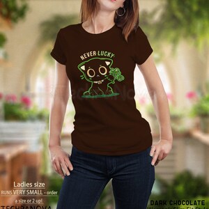 May include: A brown t-shirt with a black cat graphic holding a four-leaf clover. The text "NEVER LUCKY" is above the cat. The shirt is a women's slim fit style.