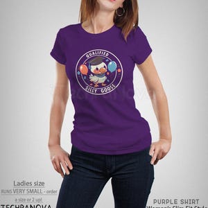 Może przedstawiać: Fioletowy T-shirt z grafiką przedstawiającą kresk&oacute;wkową gęś w czapce absolwenta trzymającą balony. Tekst "QUALIFIED SILLY GOOSE" jest nadrukowany w k&oacute;łku wok&oacute;ł gęsi.