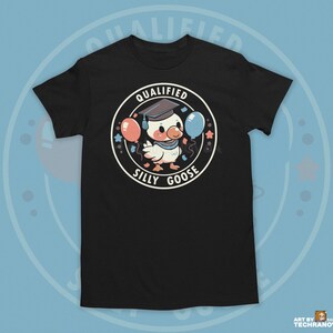 Może przedstawiać: Czarny T-shirt z kresk&oacute;wkowym gęsiem w czapce absolwenta trzymającym balony. Na koszulce nadrukowany jest napis "Qualified Silly Goose".