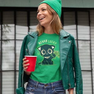 May include: A green t-shirt with a black cat illustration and the text "Never Lucky". The cat is holding a four-leaf clover. The person wearing the shirt is also wearing a green beanie and a green leather jacket.