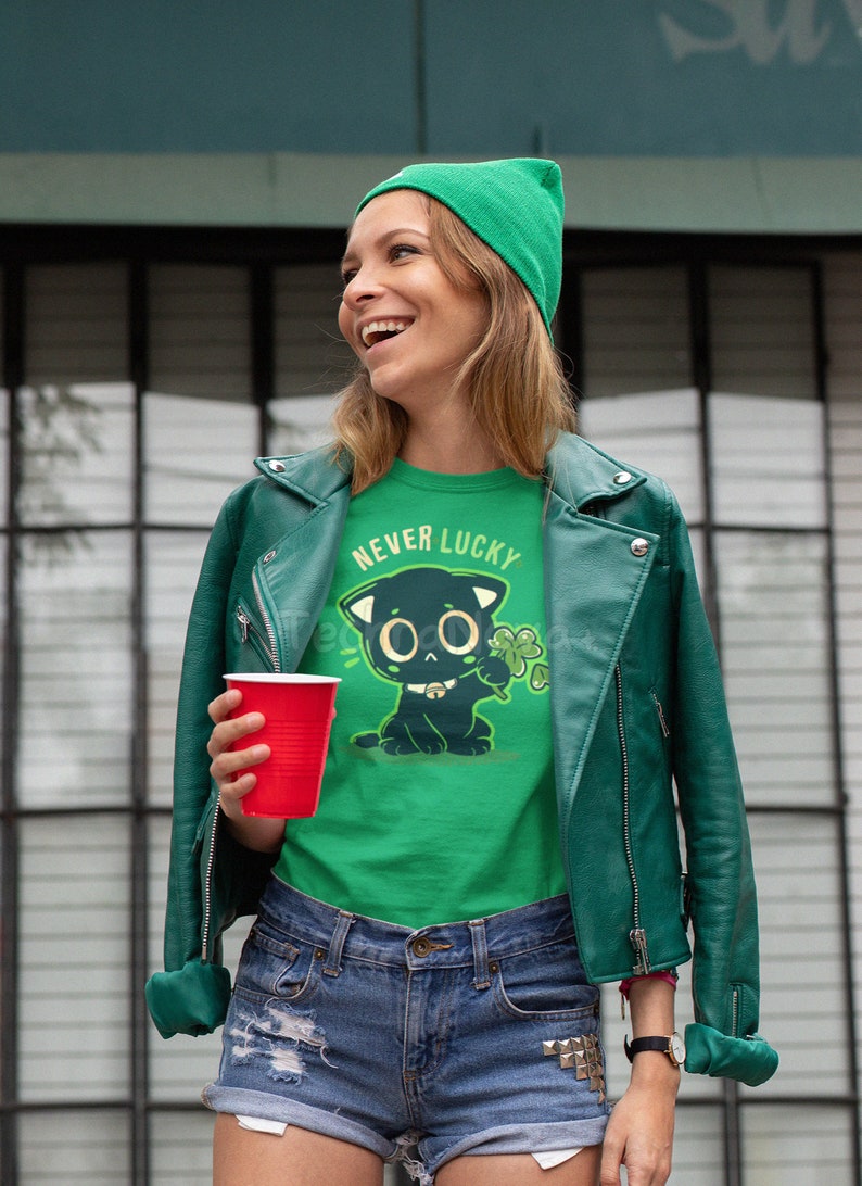 May include: A green t-shirt with a black cat illustration and the text "Never Lucky". The cat is holding a four-leaf clover. The person wearing the shirt is also wearing a green beanie and a green leather jacket.