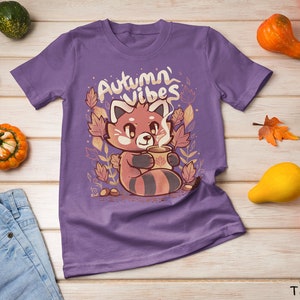 May include: Purple t-shirt with a red panda holding a cup of coffee and surrounded by autumn leaves. The text "Autumn Vibes" is printed on the shirt.